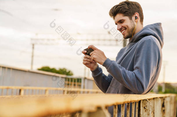 照片中，一个拿着耳机笑着的男人靠在室外栏杆上玩电子游戏