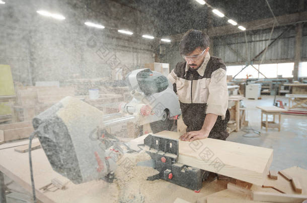 穿着制服和防护眼镜的家具制造厂的年轻工人在使用电锯时弯着腰在大木板上