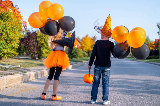 <strong>万圣</strong>节的孩子们在户外穿着<strong>嘉年华</strong>服装的后视镜兄弟姐妹。男孩和女孩戴着女巫帽,戴着南瓜糖桶.带着橙色和黑色气球的孩子们真有趣.