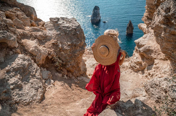 从后面看去，一个女人穿着红色的飞行服，在风中飘扬。戴着草帽，带着岩石和小船在大海的背景下走下楼梯。旅行的概念