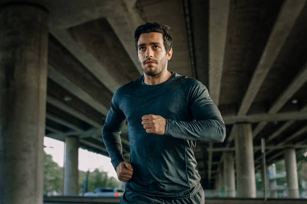 <strong>身穿运动服</strong>的运动青年男子在街上慢跑。他正在一个以汽车为背景的桥下的城市环境中跑步.