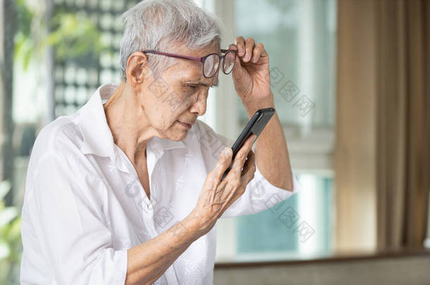 戴眼镜的亚洲老年妇女，尽量阅读短信，凝视着手机上的小短信，老年痴呆症，视力模糊，视力问题，老年痴呆症