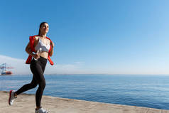 一个微笑的女运动员沿着海滨跑来跑去，欣赏美景的户外镜头。女运动员在海边慢跑