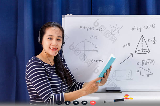亚洲女教师在家里工作，从网上教数学，从家里教学生数学。在电视大流行期间，教师在白板上写作，并在耳机网络教育课上讲话