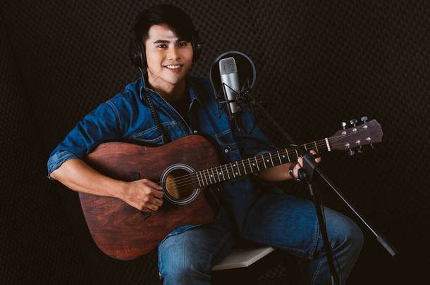 漂亮的亚洲男歌手一边弹吉他，一边用录音室麦克风和麦克风上的流行音乐盾牌在录音室里唱歌.