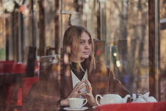 略显模糊的肖像一个迷人的女孩喝茶，通过一个咖啡馆的窗口。窗户反映了秋天的公园.