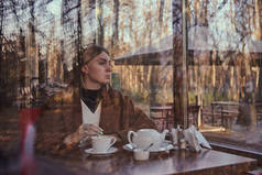 略显模糊的肖像一个迷人的女孩喝茶，通过一个咖啡馆的窗口。窗户反映了秋天的公园.