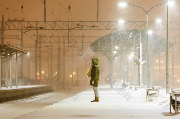 在暴风雪中的一个空荡荡的铁路站台上，一个头戴头巾的年轻女子在等着火车。运输延误。晚上天气不好，大风很大，女人们在等火车