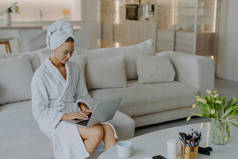 照片上漂亮的年轻女子穿着浴衣在笔记本电脑上工作，离家很远，在舒适的沙发上摆出姿势，与现代公寓的室内饮茶相映成趣。人民幸福的生活方式