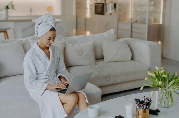 照片上漂亮的年轻女子穿着浴衣在笔记本电脑上工作，离家很远，在舒适的沙发上摆出姿势，与现代公寓的室内<strong>饮茶</strong>相映成趣。人民幸福的生活方式