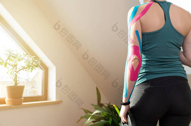 年轻健康的女人站在窗边，手里拿着运动的重物。在她的胳膊上贴上弹性运动学胶带.肌肉治疗、康复、运动和理疗。在家做运动.