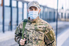 Portrait of a serious handsome young male soldier in a cap and medical mask standing in the street. 