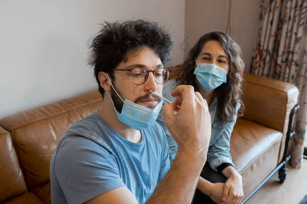 年轻的嬉皮士男子进行自我抗原检测，将鼻涕注入鼻腔以检查Covid-19的感染情况，而他的女友坐在客厅里看着他。安全保障措施.