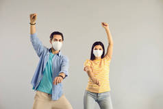 两个戴着医疗面罩的快乐年轻朋友在浅灰工作室的背景下跳舞