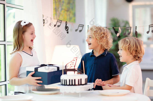 音乐和钢琴主题儿童生日派对。带着黑白蛋糕的小孩孩子吹蜡烛，打开礼物。小男孩在庆祝生日。年轻钢琴家的活动。节日装饰.