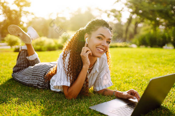 年轻的自由职业女子坐在绿草上，手持<strong>笔记本</strong>电脑。网上教育。快乐的女人，带着无线耳机在<strong>笔记本</strong>电脑上呼叫，用网络摄像头交谈，开视频会议.