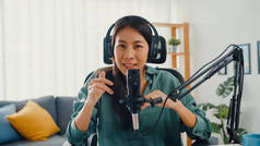 快乐的亚洲女孩带着耳机和话筒录制了一个播客，看着摄像机说话，然后在房间里休息。女播音员从她的家庭演播室制作音频播客。.