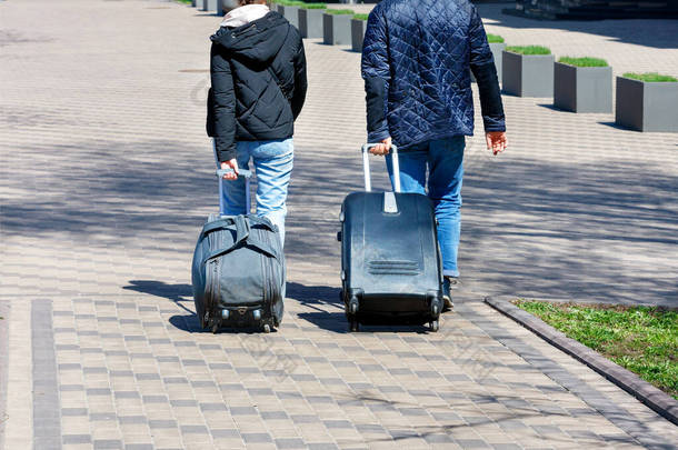 一对年轻夫妇，一个男人和一个女人，走在鹅卵石铺就的人行道上，在阳光灿烂的日子里旅行，背着旅行袋和车轮提着手提箱。复制空间.