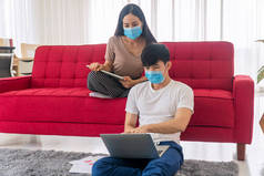 在检疫验尸官大流行期间，戴口罩保护眼罩的年轻夫妇在沙发上坐着工作时使用笔记本电脑。生活方式爆发概念