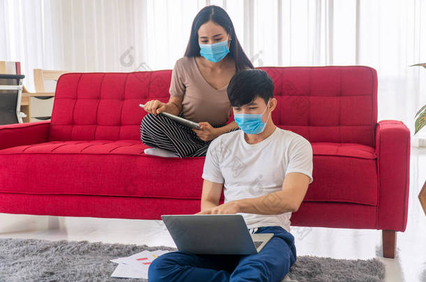 在检疫验尸官大流行期间，戴口罩保护眼罩的年轻夫妇在沙发上坐着工作时使用笔记本电脑。生活方式爆发概念