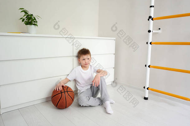 一个学龄前<strong>男孩</strong>坐在他房间的地板上，抱着一个关于体育活动的<strong>篮球</strong>梦。在家里隔离。一个孩子打<strong>篮球</strong>的肖像.
