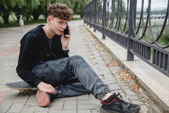 一个身穿黑色连帽衫、卷曲头发的欧洲少年坐在滑板上的小巷里，一边打电话一边聊天