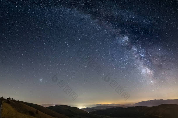 意大利圣斯蒂凡诺、阿布鲁佐和罗卡 · 卡拉索上空的全景<strong>夜空</strong>。<strong>银河系</strong>的弧形和星空照亮了村庄独特的山水景观.木星行星可见.