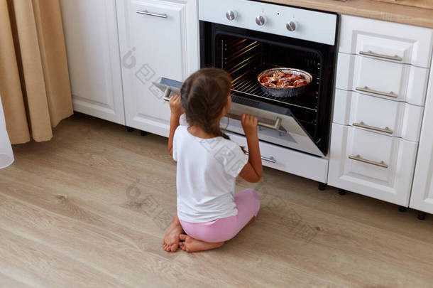 在烤箱附近等待烤羊角面包、松饼或纸杯蛋糕的小女孩，坐在地板上看着烤箱里面的小女孩，穿着白色休闲衫的小女孩.