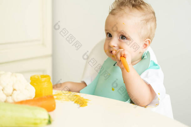 滑稽的小男孩，穿着围裙，坐在儿童椅子上，用勺子吃蔬菜泥。小孩自己学习吃东西.