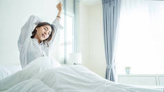 身穿睡衣的亚洲美女早上醒来，心情很愉快。迷人的年轻女子笑着、快乐着、放松着，然后从床上躺在家里的床上舒展身体.;