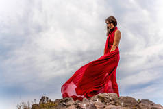 一个穿着红色衣服的女人站在暴风雨的天空之上，裙子飘扬，面料在风中飘扬.