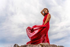一个穿着红色衣服的女人站在暴风雨的天空之上，裙子飘扬，面料在风中飘扬.