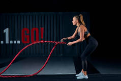 强壮的肌肉发达的女人用战斗绳锻炼。在体育馆里穿着时髦运动服的漂亮女人的照片。侧视图.
