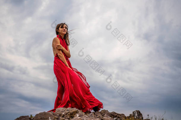 一个穿着红色衣服的女人站在暴风雨的天空之上，裙子飘扬，面料在风中飘扬.