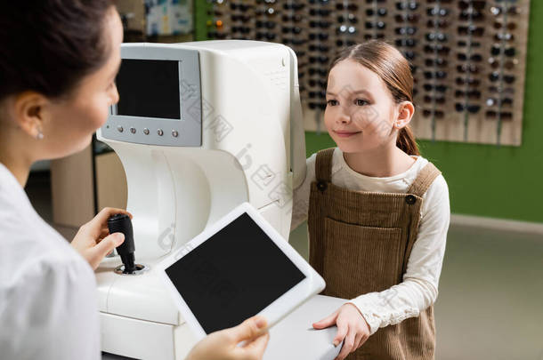 光学商店里，模糊的眼科医生拿着空白屏幕的数码平板电脑，接近阳性女孩和视力检查仪