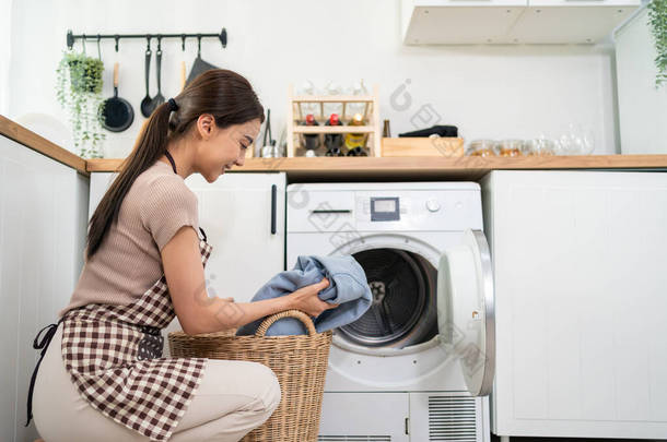 亚洲美女把脏衣服放在洗衣机里.迷人的女孩穿着围裙坐在地板上，高高兴兴地在家里的洗衣机里洗衣服。家政服务的概念.