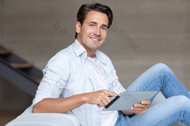 在家里的沙发上挂着数字平板电脑，看着网上购物和上网的相机，面带微笑的英俊男子.