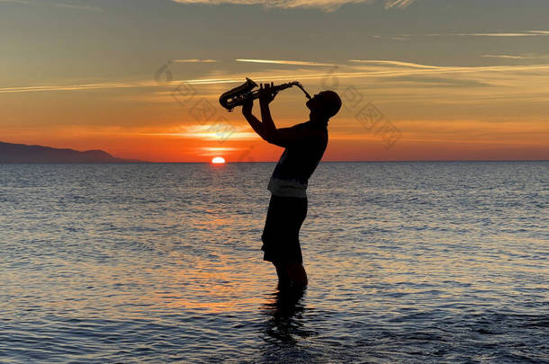 年轻的萨克斯风手脚踏着海水站在那里，手里拿着萨克斯风，望着日落。美丽的落日在海上，天空。音乐家，吹奏萨克斯风，跳舞，玩乐，