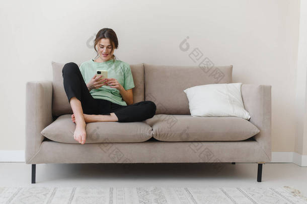 照片中快乐而乐观的年轻女性坐在室内，购物或社交媒体在沙发上使用手机.