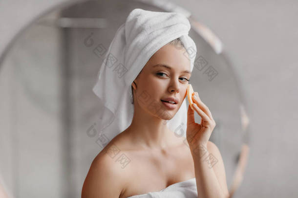 漂亮女士用化妆品海绵享受日常的面部肌肤护理，早上站在浴室里