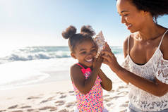 美丽的年轻母亲在海滩边，在快乐的女儿耳边抱着贝壳，听着大海的声音。可爱的小女孩听到驾驶舱地狱与妈妈在热带海滩。笑着和听着的女人在一起的小孩