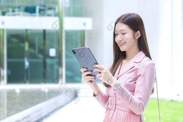 美丽的年轻亚洲职业女性留着长发，在办公室前的花园里微笑着，手里拿着平板电脑与办公室里的客户见面，从任何概念出发工作.