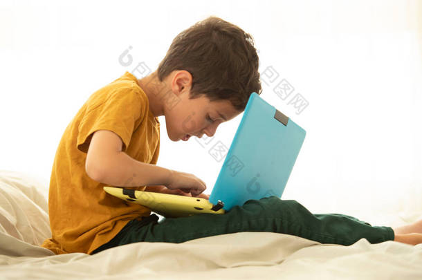 孩子们坐在床上，带着一台黄色和蓝色的笔记本电脑，专心致志地在键盘上打字。虚拟游戏、网上教育、远程学习、学习、网络安全、健康姿态、弯腰的概念.