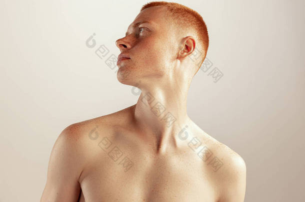 年轻红<strong>头发男子</strong>的画像，穿着无袖上衣，在灰色摄影棚的背景上看起来是孤立无援的。男性健康、生活方式、美感、身体和皮肤护理的概念。有雀斑的模型