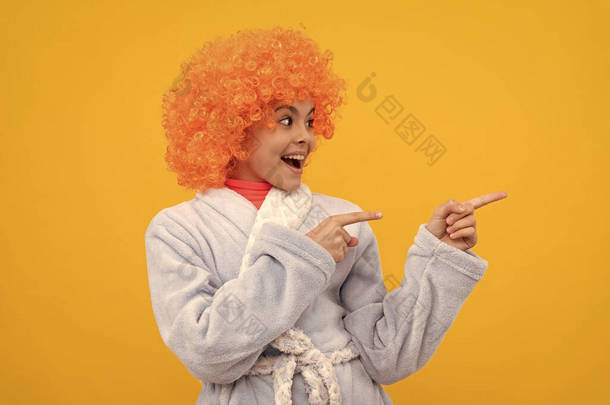快乐的孩子穿着华丽的橙色假发，头戴家用浴衣，指指点点复印空间，做广告.