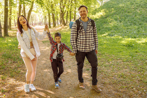 快乐快乐的年轻母亲、父亲和小男孩手牵着手在绿林中漫步。在公园里散步