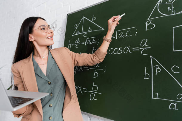 课堂上，老师在接近黑板的地方拿着<strong>笔记本</strong>电脑，<strong>上面</strong>有数学公式