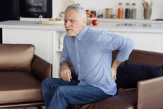 老年人腰痛的侧视图照片，坐在舒适的矫形外科扶手椅上，姿势不正确。慢性损伤、肌肉炎症和背痛概念