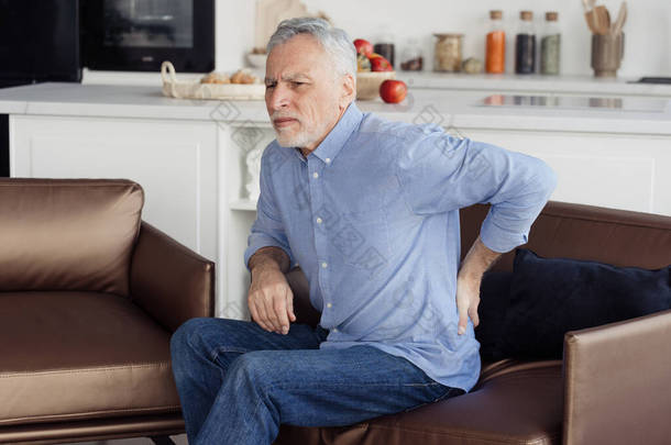 老年人腰痛的侧视图照片，坐在舒适的矫形外科扶手椅上，姿势不正确。<strong>慢性</strong>损伤、肌肉<strong>炎症</strong>和背痛概念