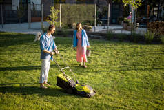 男人用割草机割草，夏天和妻子一起在乡间别墅的后院度过。夏天的傍晚，年轻的家人照料花园